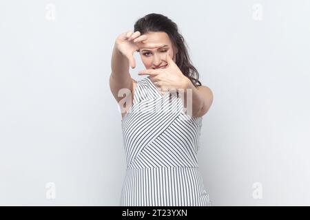 Portrait of woman searches perfect angle takes photo, makes frame gesture as if photographing with camera, smiles positively, wearing striped dress. Indoor studio shot isolated on gray background. Stock Photo