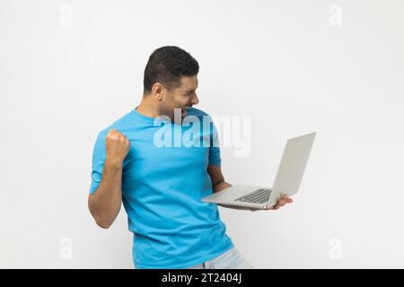 Joyful cheerful unshaven man in blue T- shirt standing holding laptop in hands, clenches fist, celebrating success, finishing project till deadline. Indoor studio shot isolated on gray background. Stock Photo