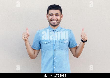 Portrait of extremely happy joyful cheerful man wearing denim shirt looking at camera with toothy smile, showing thumbs up, approved. Indoor studio shot isolated on gray background. Stock Photo