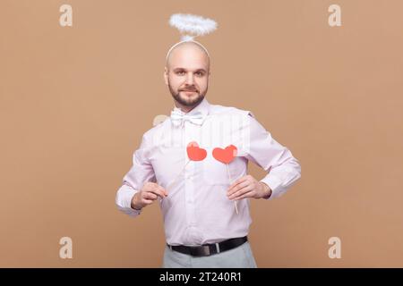 Portrait of cute friendly bald bearded man with nimb over head, holding little red hearts in front of his chest, wearing light pink shirt and bow tie. Indoor studio shot isolated on brown background. Stock Photo