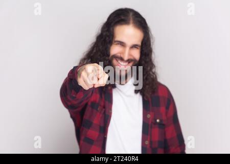You got it. Happy bearded man with long curly hair in checkered red shirt points at camera with index fingers, picks someone smiles broadly. Indoor studio shot isolated on gray background. Stock Photo