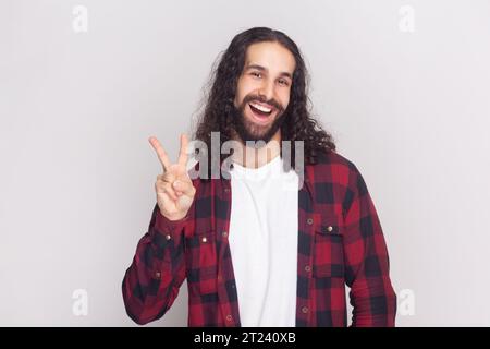 Portrait of optimistic glad bearded man with long curly hair in checkered red shirt with positive facial expression, shows V sign or victory gesture. Indoor studio shot isolated on gray background. Stock Photo