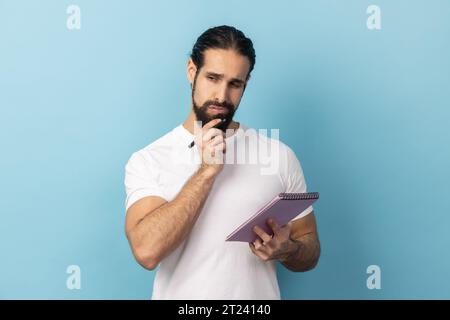 Portrait of thoughtful man with beard wearing white T-shirt holding paper notebook, having thoughtful facial expression, planning. Indoor studio shot isolated on blue background. Stock Photo