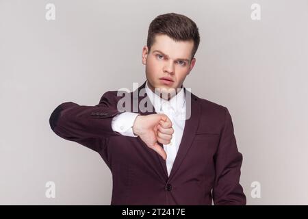 Portrait of handsome man shows disapproval sign, keeps thumb down, expresses dislike, frowns face in discontent, wearing violet suit and white shirt. Indoor studio shot isolated on grey background. Stock Photo