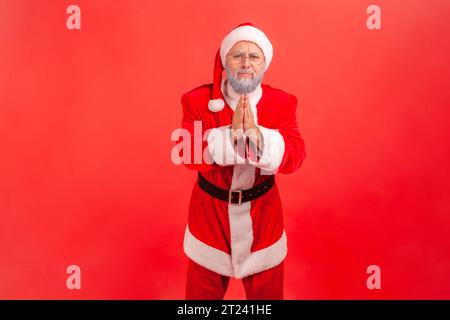 Forgive me or give me one chance more. Elderly man with gray beard wearing santa claus costume standing with palm hands, looking and begging. Indoor studio shot isolated on red background. Stock Photo