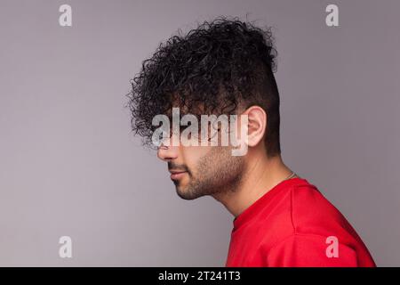 Side view portrait of attractive handsome young adult bearded man with curly hair, wearing red jumper, looking ahead, showing his hairdo. Indoor studio shot isolated on gray background. Stock Photo