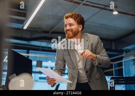 Smiling businessman holding documents and looks away while standing in office Stock Photo