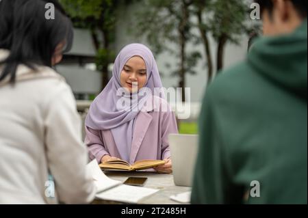 A young Asian-Muslim female college student is reading a book or doing homework with her friends in a campus park. Education and diverse students conc Stock Photo
