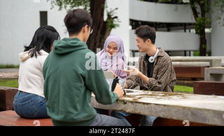 A cheerful young Asian-Muslim female college student is doing homework or a group project with her friends at a table in a campus park. Diverse studen Stock Photo
