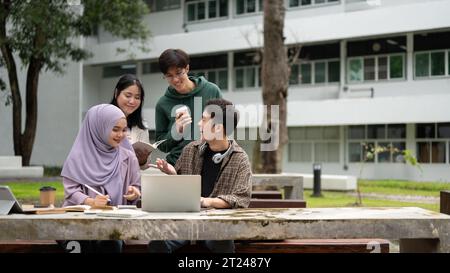 A group of diverse young college students is discussing project details, using a laptop, and working on a group project at a table in a campus park to Stock Photo