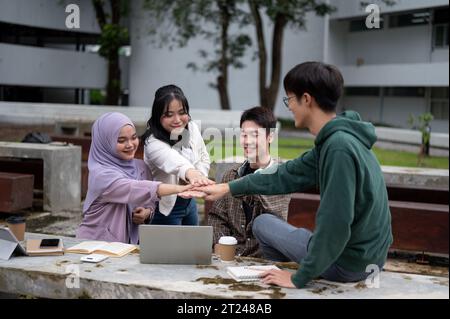 A group of cheerful, diverse Asian university students is putting their hands together at a table in a campus park, encouraging and supporting each ot Stock Photo