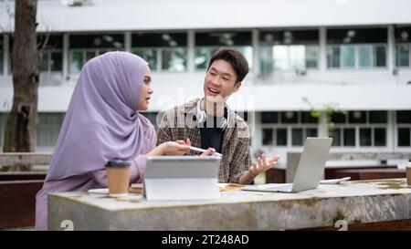 A smart and cheerful young Asian male university student is talking and helping his female Muslim friend with math at a table in the park. Friendship Stock Photo