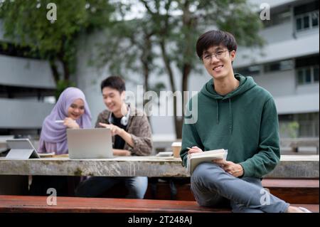 A smart and handsome young Asian male university student in a green hoodie and eyeglasses is sitting at a bench in a campus park with his friends. Edu Stock Photo