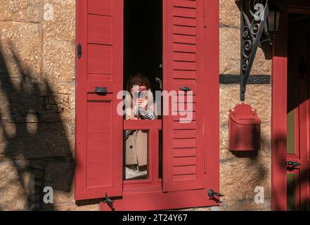 10152023,Edremit,Balikesir,Turkey,Woman tasting wine behind the window with red shutters in Adatepe village Stock Photo