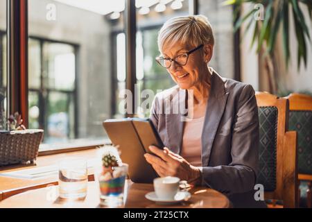 Mature woman is sitting in cafe and relaxing. She is drinking coffee and using digital tablet. Stock Photo