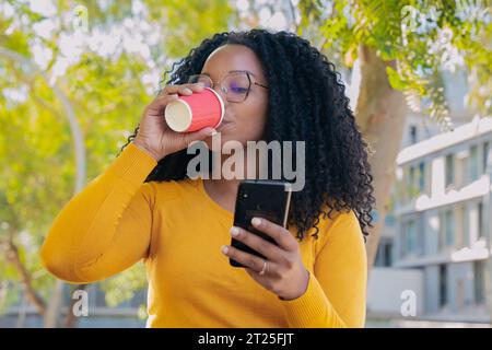 Young smiling black woman at mobile phone while drinking coffee. Stock Photo