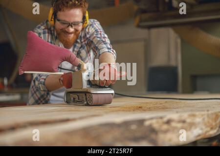 Close up of smiling bearded male wood worker using electric sander for wood. Carpentry workshop Stock Photo