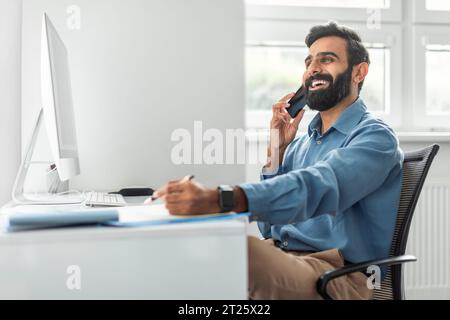 Positive thoughtful indian businessman talking on cellphone and taking notes while working on computer in office Stock Photo