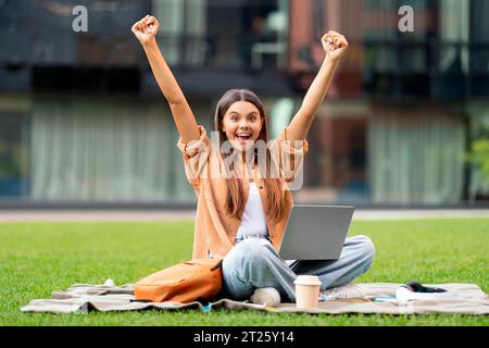 Emotional woman student with laptop on lap raising hands up Stock Photo