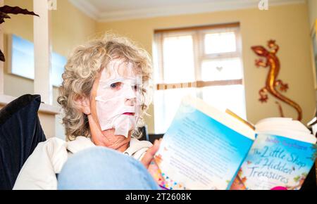 Mature woman reading a book at home wearing a face pack to try and get rid of unwanted wrinkles and keep her skin smooth Stock Photo