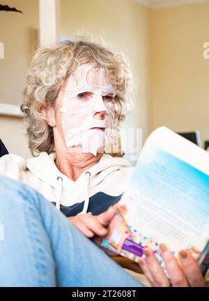 Mature woman reading a book at home wearing a face pack to try and get rid of unwanted wrinkles and keep her skin smooth Stock Photo