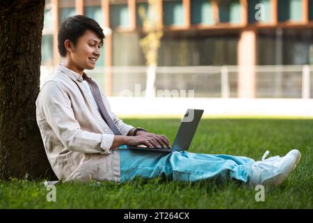 Concentrated asian man sitting under tree, working on his laptop Stock Photo
