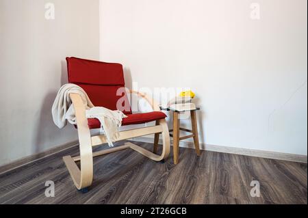 Red wooden modern armchair with opened book and yellow flowers on the small table near it. Decor, style Stock Photo