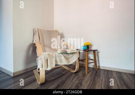 Wooden modern armchair cowered with cotton plaid, opened book on it and yellow flowers, blue cup on the small table near it. Decor, style Stock Photo