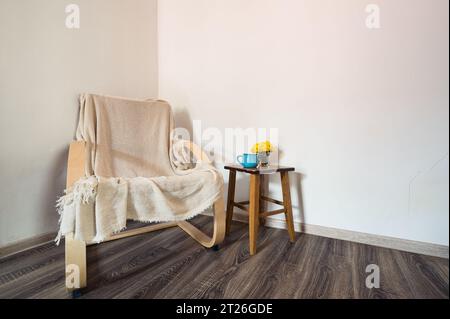 Wooden modern armchair covered with white plaid, vase of yellow flowers and blue cup on the small table near it. Decor, style Stock Photo