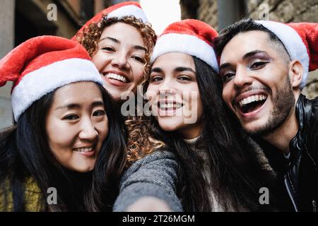 Diverse group of friends having fun celebrating Christmas on city street - Young people taking selfie picture wearing Santa Claus hats during holidays Stock Photo