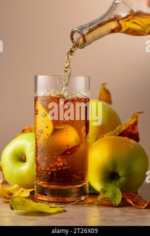 Apple juice is poured from a bottle into a glass. On a ceramic table ripe apples and dried-up leaves. Stock Photo