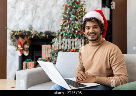 Portrait of young man at home, hispanic smiling and looking at camera, sitting on sofa near Christmas tree and presents, using laptop for remote work, and sending New Year's greetings to family . Stock Photo