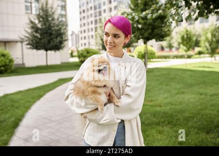 happy purple-haired woman holding fluffy dog in hands and looking away on street, city walk Stock Photo