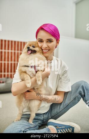 purple-haired woman sitting on floor and embracing pomeranian spitz, affection of dog and pet sitter Stock Photo