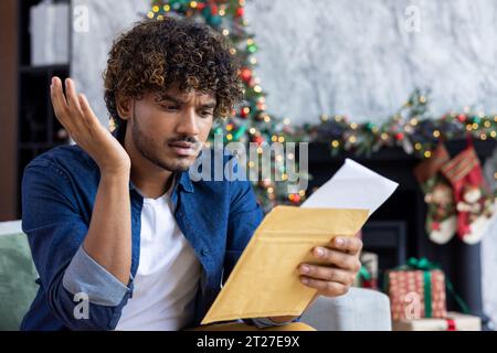 Man received postal envelope letter with bad news, upset disappointed hispanic man reading notification sitting at home in living room on sofa, christmas and new year celebration. Stock Photo