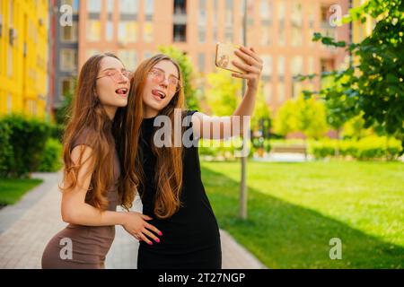Funny two young twin sisters with cool sunglasses using Social media and taking a Selfie With smartphone in with a backdrop of colorful buildings Stock Photo