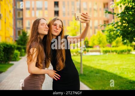 Funny two young twin sisters with cool sunglasses using Social media and taking a Selfie With smartphone in with a backdrop of colorful buildings Stock Photo