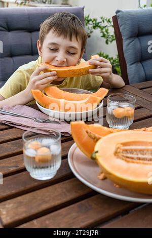 Cute preteen boy with close eyes sitting at table and eating sliced fresh musk melon fruit on plate with melon spheres in iced beverage against blurre Stock Photo