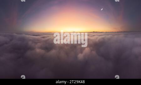 Breathtaking aerial view over white thick and soft clouds floating against mountain peaks at colorful sundown in evening time with moon Stock Photo