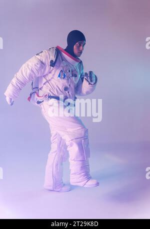 Full body of male ethnic astronaut in spacesuit and backpack with dark hood marching with raised leg over purple background Stock Photo