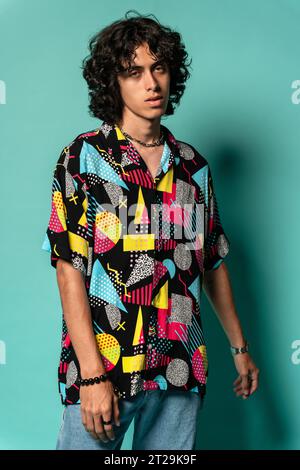 Confident young Latin American male in colorful shirt dreamy eyes looking at camera while standing with shadow against turquoise background in light Stock Photo