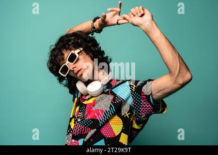 Young Latin American male in sunglasses looking at camera while standing with headphones on neck and holding fingers over head at turquoise background Stock Photo