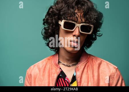 Funky young Latin American dancer in sunglasses curly hair looking at camera while standing in pink jacket against turquoise background Stock Photo