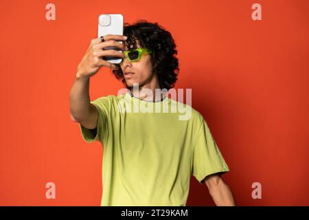 Smiling young Latin American male with dark hair looking at screen of mobile phone while standing with sunglasses during selfie at red background Stock Photo