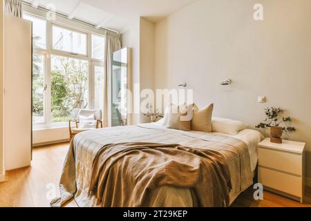 Comfortable bed with pillows and blankets arranged by large bright window in modern bedroom with white walls Stock Photo