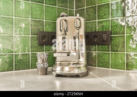 Stainless steel coffee maker with cups placed on counter in front of green tiled wall in kitchen Stock Photo