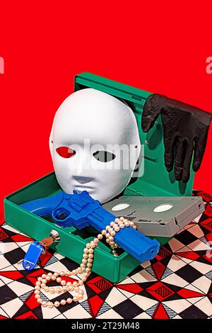 High angle of opened secret box with mask, gloves, toy gun, necklace and video tape placed on table against red wall Stock Photo