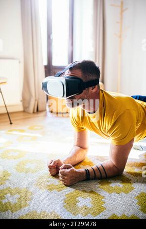 Fit bearded guy in yellow T shirt in VR glasses doing exercise on rug in light cozy room Stock Photo