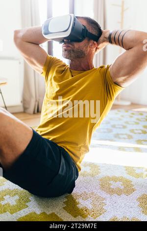 Muscular bearded male in VR headset doing abs on carpet while working out in room at home on sunny day Stock Photo