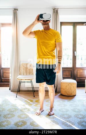 Full body of barefoot man in activewear looking through VR glasses while standing in cozy room in sunlight Stock Photo